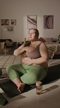 Vertical video of plus-size young adult Caucasian woman sitting on yoga mat with closed eyes, practicing deep breathing and meditation. Clip is ideal for body positivity and wellness campaigns