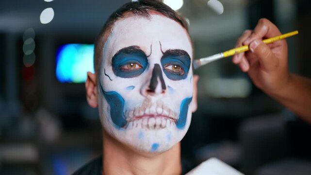Close-up of man receiving traditional Day of the Dead skull makeup application. Artist uses brush to add blue and black details to white painted face with skeleton teeth design.
