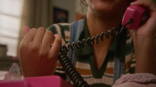 African American teenage girl talking on pink retro landline telephone while twisting coiled cord in home bedroom. Suitable for nostalgic marketing campaigns or youth lifestyle content