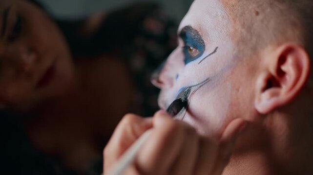 Close-up of makeup artist carefully applying traditional Day of Dead or Halloween face paint on young man. White base with blue eye shadows and black skull details create dramatic sugar skull effect.