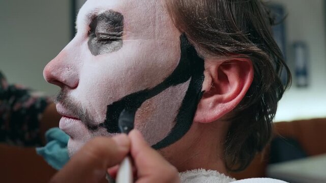 Close-up of professional makeup artist applying traditional Day of the Dead or Halloween face paint on man. White base with black geometric patterns being painted on cheek and jawline using brush.