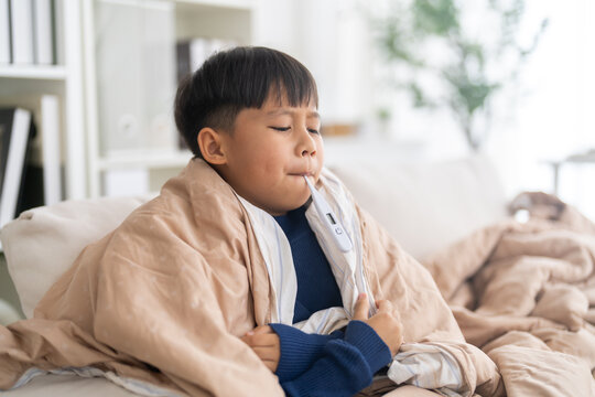 Asian boy showing fever and headache holding thermometer in mouth, flu symptoms in children, covid-19 illness, home healthcare, hospital visit, influenza care, caregiving and wellness, sick at home
