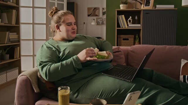 Plus-size Caucasian young adult woman lying on sofa, eating sandwich while watching video on laptop. Perfect for body positivity, remote work, and food delivery service advertisements