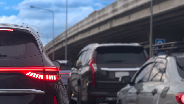 Low angle shot showing the rear of a black car stuck in dense traffic During the day. Turn on brake light. Vehicles are lined up on the highway road beneath a massive concrete overpass structure.