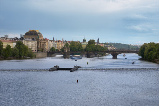 View of the wide Vltava River in Prague with a weir in the foreground and a historic stone bridge in the distance under an overcast sky.