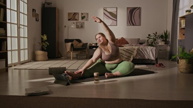 Plus-size Caucasian young adult woman stretching on yoga mat at home while watching online tutorial on tablet. Video is perfect for body positivity marketing and healthy lifestyle advertisements