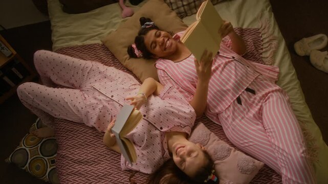 Two multiethnic teenage girls lying on bed in pajamas, reading books and talking during sleepover. Ideal for youth lifestyle brands, bedding advertisements, or educational campaigns