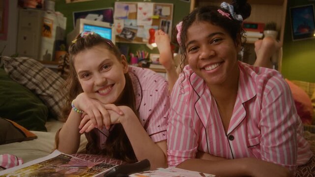 Portrait of Caucasian girl and African American teenager lying on bed in pajamas and smiling at camera, enjoying slumber party. Footage is ideal for youth lifestyle marketing and diversity campaigns