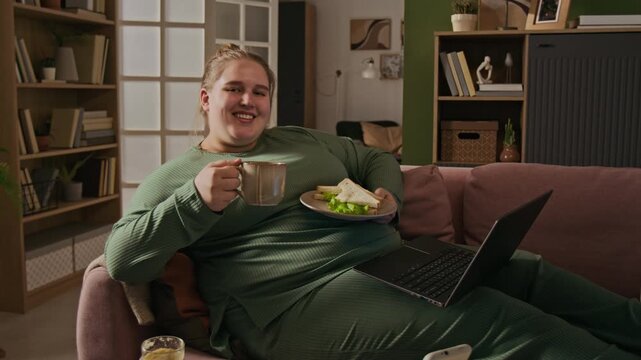 Portrait of smiling plus-size young adult Caucasian woman lying on sofa with laptop, holding cup and sandwich. Clip is perfect for body positivity, remote work, and lifestyle marketing campaigns