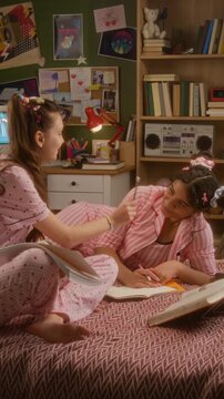 Vertical of diverse teenage girls in pajamas sitting on bed, writing in notebooks, talking and laughing while doing homework. Youth lifestyle, education, or sleepwear brand marketing campaigns