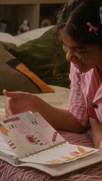 Vertical of African American teenage girl lying on bed and flipping through decorated scrapbook diary or journal. Youth lifestyle brands, mental health apps, or creative hobby promotions