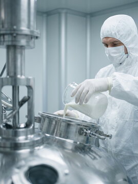 Scientist in protective suit pouring liquid into large steel mixer in sterile environment, pharmaceutical production.