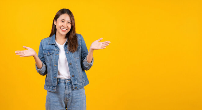 Smiling young asian woman in casual outfit presenting with open hands on bright yellow background, cheerful confident model jeans for lifestyle and advertising concepts