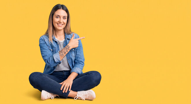 Smiling young woman sitting cross-legged on bright yellow background pointing to the side in casual outfit with tattoo and relaxed confident pose