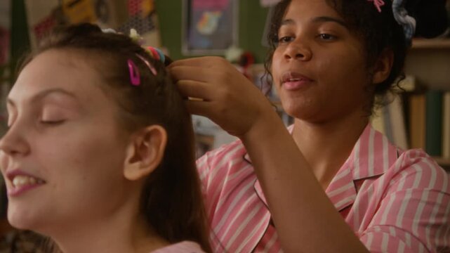 African American teenage girl styling hair of Caucasian friend during slumber party while chatting and gossiping. Footage suitable for high school youth lifestyle, friendship, and hair care marketing