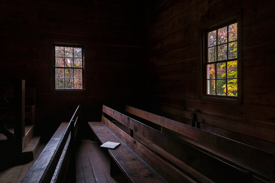 Dark interior of a church with a bible on a pew and fall foliage visible through the windows