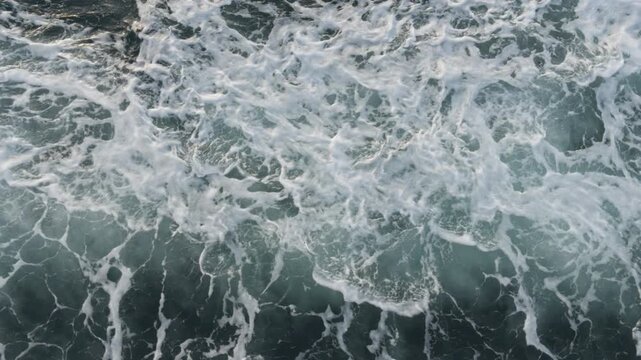 Static high-angle shot of ocean water churning violently, creating thick white foam, bubbles, and sea spray in the wake of a moving vessel on a bright day.