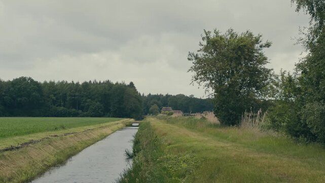 Peaceful Dutch rural landscape featuring a canal winding through farmland, a dike along the waterway and forest bordering green pastures. A calm countryside scene beneath an overcast sky.
