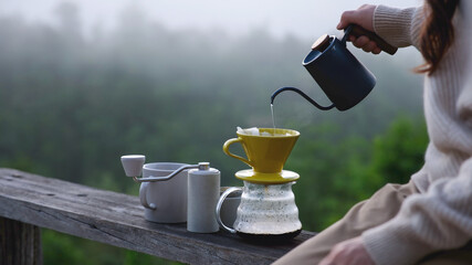 Naklejka premium Closeup of a woman making drip coffee on wooden terrace with a mountain view on foggy morning