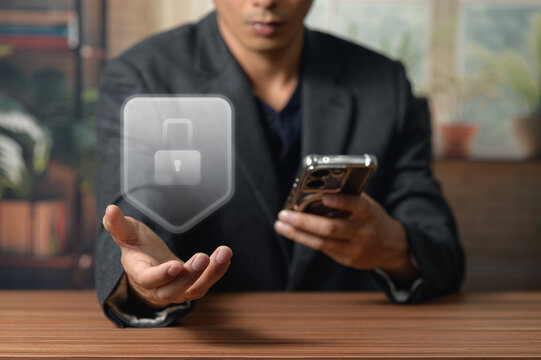 Man in dark blazer holding smartphone with holographic lock symbol at desk in office