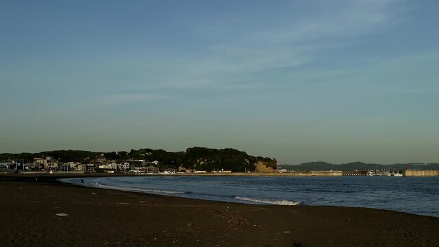 Scenic beach view in Kamakura, Japan, with gentle ocean waves, dark sand, blue sky and distant island silhouette. Peaceful coastal travel scene for seaside tourism and relaxation themes.
