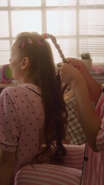 Vertical of African American teenager braiding hair of Caucasian friend during pajama party while gossiping. Footage is perfect for beauty campaigns or youth lifestyle marketing