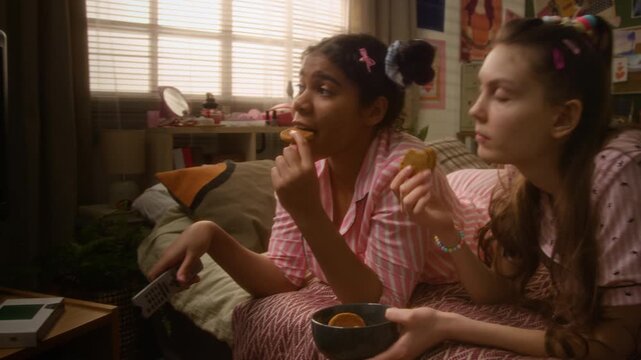 African American and Caucasian teenage girlfriends lying on bed, eating cookies, watching fun movie retro television and laughing. Streaming service advertisements or youth lifestyle campaigns