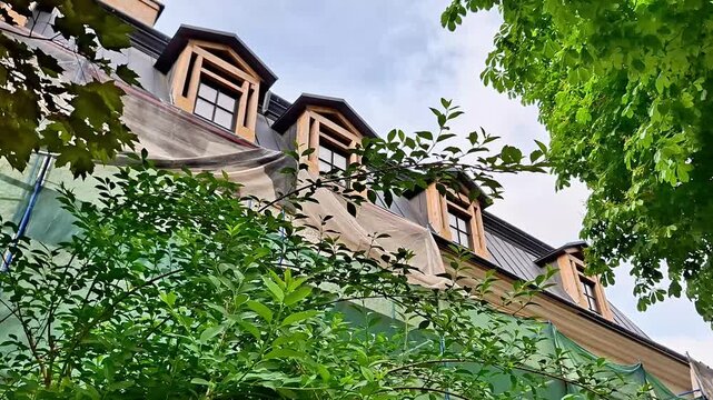 Low angle view of a charming mansard roof with classic dormer windows framed by lush green trees and shrubs. A peaceful architectural scene blending historic design with vibrant nature