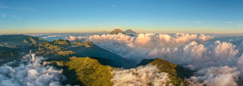 Aerial view of the majestic Mount Bismo peaks rising above a sea of clouds at sunrise with lush green slopes and distant volcanic summits in Kecamatan Kejajar, Jawa Tengah, Indonesia.