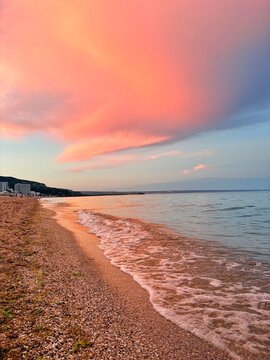 Stunning coastal landscape featuring dramatic pink and orange sunset clouds reflecting on the calm sea water and sandy shoreline.
