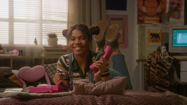 Portrait of smiling African American teenager lying on bed and holding pink retro telephone receiver, vintage computer in babckground. Nostalgic marketing campaigns or youth lifestyle commercials