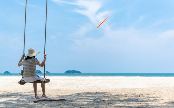 Summer sandy beach holiday vacation with young girl relaxing sitting on swing enjoying tropical ocean view, beautiful blue sunny sky on sea shore