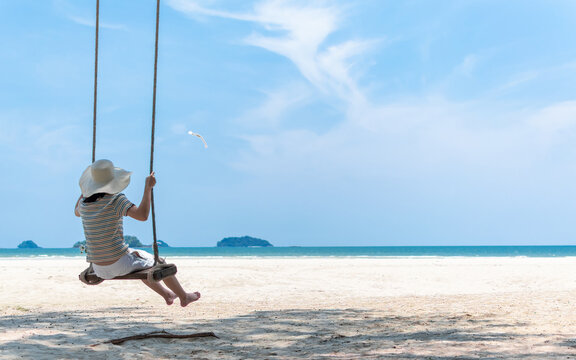 Summer sandy beach holiday vacation with young girl relaxing sitting on swing enjoying tropical ocean view, beautiful blue sunny sky on sea shore