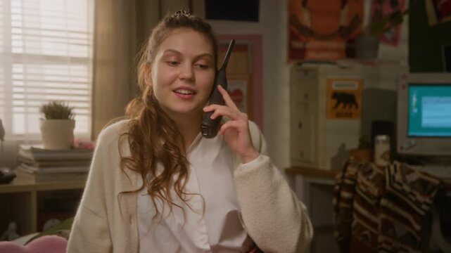 Young Caucasian girl talking on retro cordless phone in 90s themed bedroom, smiling, flirting and playing with hair. Clip suitable for nostalgia marketing campaigns and retro technology commercials
