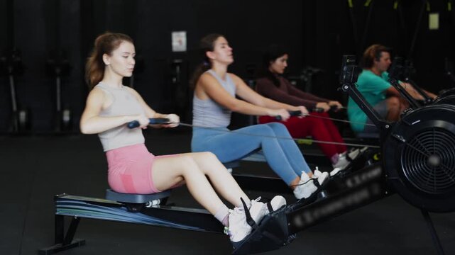 Young woman with sports leggings and a top does a cardio workout on a rowing machine. Crossfit group training participants perform a circuit training session for endurance.