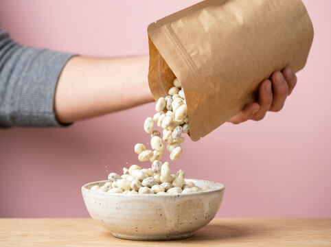 Pouring white beans from a brown paper bag into a ceramic bowl, healthy plant-based cooking ingredients, raw organic navy beans on pink background