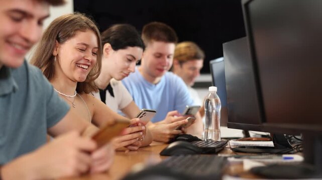 Cheerful young female student studying together with coursemates in computer workshop, actively using mobile phone to access online learning materials or participating in group discussions