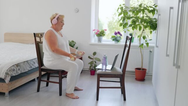 Active elderly female doing fitness, bicep curls with yellow dumbbells in bright room