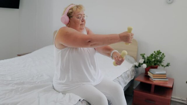 Overweight elderly lady doing dumbbell raises sitting on bed at home fitness at home