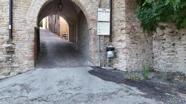 Arco in pietra che introduce a una stretta via lastricata tra edifici storici in mattoni, con lampada centrale e parete laterale coperta da vegetazione. Scena urbana antica caratteristica