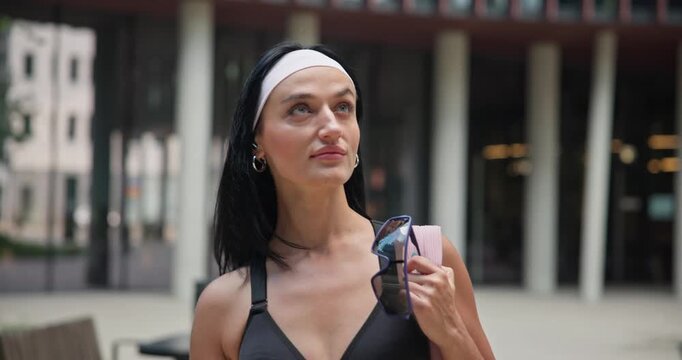 A young beautiful female athlete in sportswear and a backpack goes for a workout in an urban park among high-rise buildings