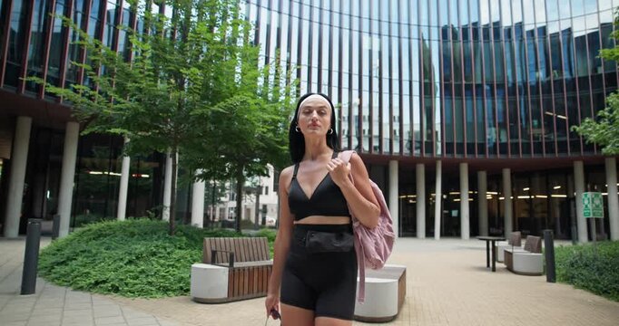 A young beautiful female athlete in sportswear and a backpack goes for a workout in an urban park among high-rise buildings