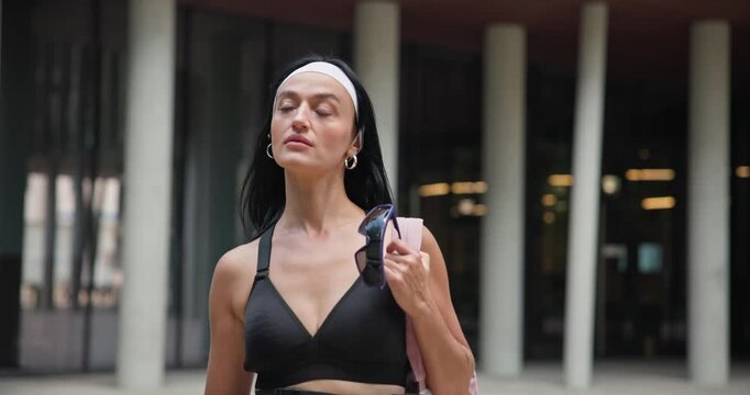 A young beautiful female athlete in sportswear and a backpack goes for a workout in an urban park among high-rise buildings