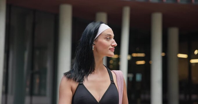 A young beautiful female athlete in sportswear and a backpack goes for a workout in an urban park among high-rise buildings