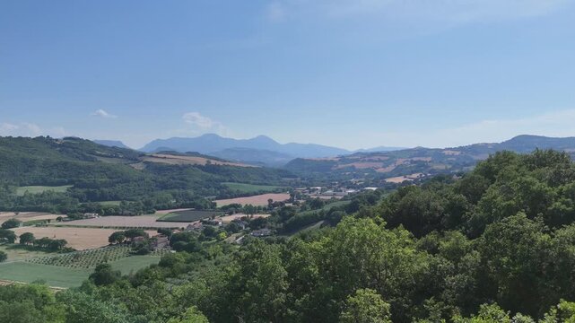Paesaggio rurale con colline verdi, campi coltivati, alberi e montagne in lontananza sotto cielo azzurro limpido. Scena naturale ampia, luminosa e tranquilla