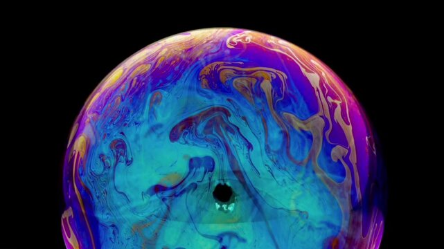 A close-up of a spherical soap bubble against a pitch-black background. An iridescent effect creates a dynamic blend of colors, ranging from cyan blue, purple, and pink to golden yellow. 
