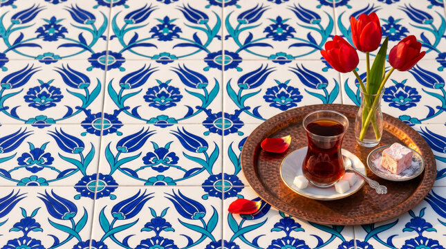 Turkish tulip-shaped tea glass with amber brew, sugar cubes, copper tray, red tulips and lokum on Iznik tiles, copy space. Tea advertising concept