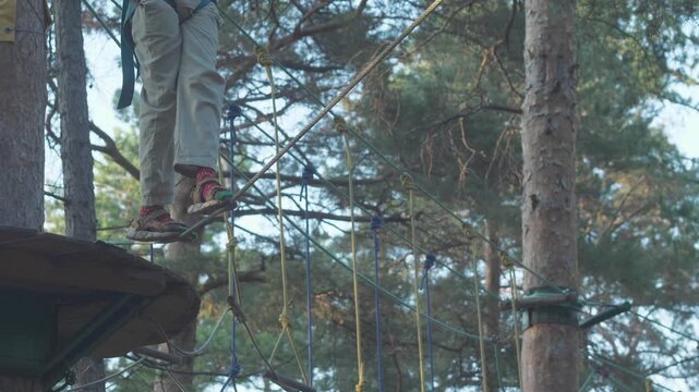 A teenager in a safety harness starts moving from a wooden platform in a rope park. Close-up of the legs of a guy carefully stepping on a thin stretched cable at a height.