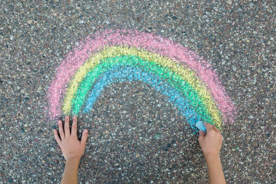 Little child girl hand fingers holding blue chalk and drawing colorful rainbow on dark gray asphalt background in summer day. Closeup. Point of view shot. Top down view.