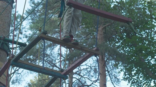 A teenager in a safety harness passes a difficult high obstacle made of wooden frames. A guy balances on swaying bars in an amusement rope park against the background of trees.